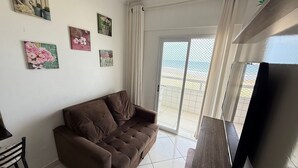 A living room with a brown sofa, a TV, and a sliding glass door leading to a balcony. - Foot in the sand at Maracanã beach II (Praia Grande)