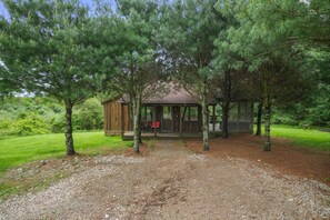 A wooden cabin with a covered porch, surrounded by trees and a gravel path.