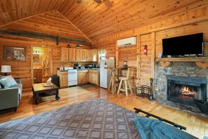 A cozy cabin interior with wooden walls, a stone fireplace, a kitchen area, and a living room with a sofa and a coffee table.