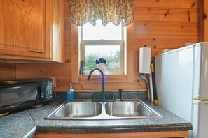 A kitchen with wooden cabinets, a double sink, a microwave, a refrigerator, and a window with a view of greenery.