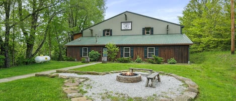 A rustic building with a green roof, a fire pit in the center, and a gravel path leading to the entrance.