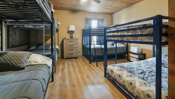 Una camera in dormitorio con letti a castello, soffitto in legno, ventilatore a soffitto, comodino con lampada e finestra con tende.