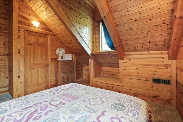 Un dormitorio de cabaña de madera con techo inclinado, una cama con colcha estampada, un ventilador pequeño y una ventana con cortina azul.