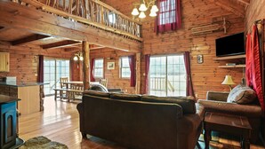 A rustic cabin interior with a wooden ceiling, a dining area, a sofa, a television, and a stone fireplace. - Pioneer Lodge | Hocking Hills, Sleeps 15, Hot Tub (Logan)