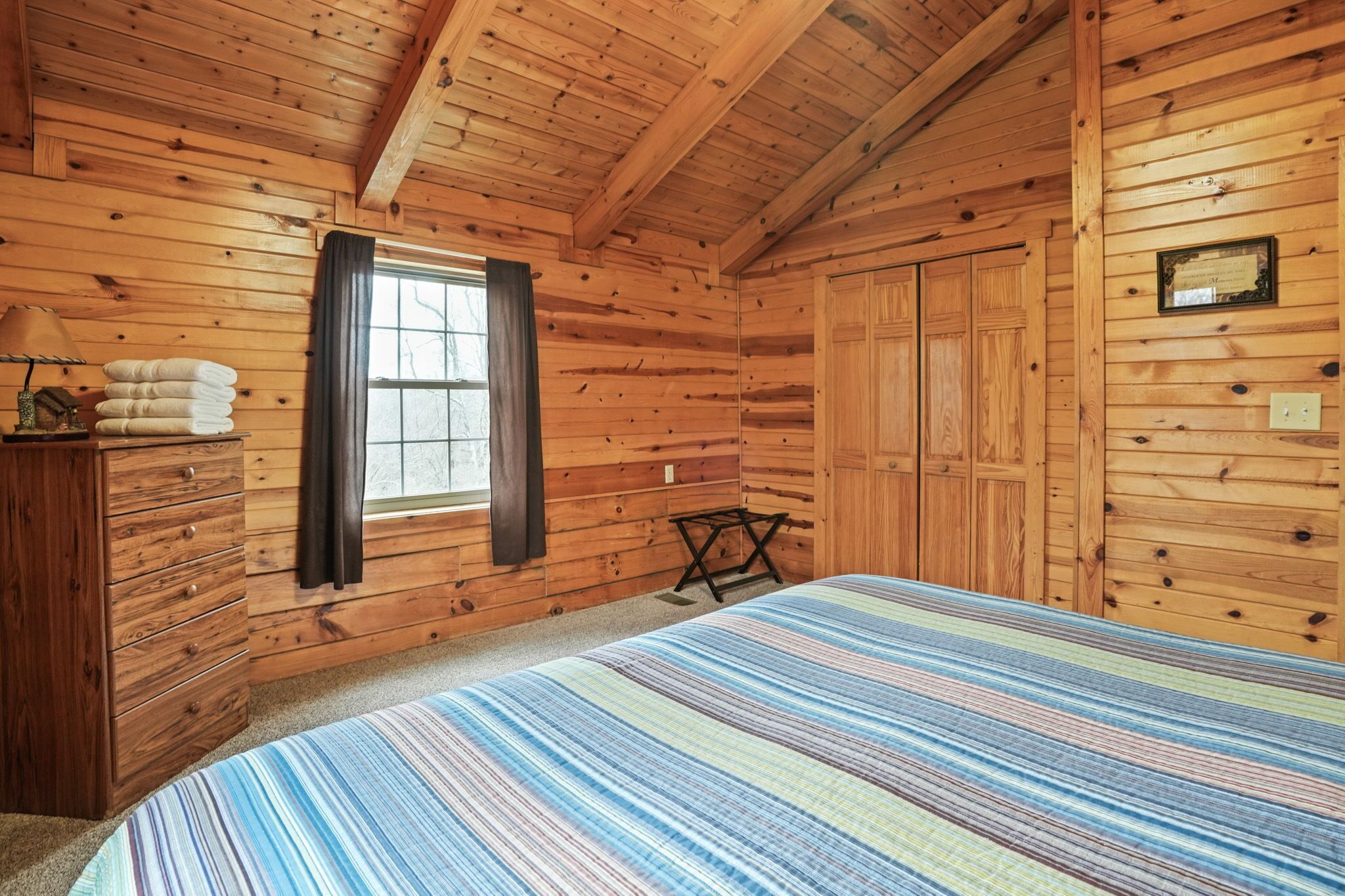 A cozy log cabin room with a bed, a window with curtains, a wooden dresser, and a framed certificate on the wall.