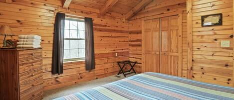 A cozy log cabin room with a bed, a window with curtains, a wooden dresser, and a framed certificate on the wall.