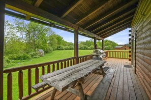 Una terraza de madera con mesa de picnic y parrilla, con vista a un patio césped y un edificio pequeño.