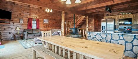 Interior de una cabaña de madera con una larga mesa de comedor, bancos, una pared de acento de piedra, una zona de cocina y una escalera.