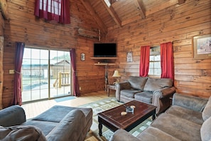 Interior de una cabaña de madera con sofá, sillones, una mesa de centro, televisor y un ventanal con cortinas.