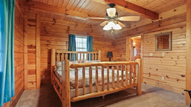 A wooden cabin bedroom with a wooden bed, a ceiling fan, and a window with curtains. - Pioneer Lodge | Hocking Hills, Sleeps 15, Hot Tub (Logan)