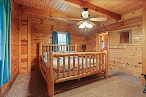 Un dormitorio de cabaña de madera con cama de madera, ventilador de techo y ventana con cortinas.