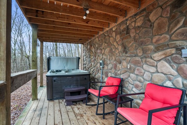 A covered outdoor area with a stone wall, a hot tub, and red chairs. - Hide-A-Way Tiny House | Hocking Hills, Hot Tub (Logan)