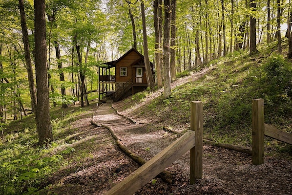 A cabin in a forest with a wooden deck and a dirt path leading to it.