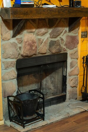 A stone fireplace with a metal grate, a black metal stand, and a wooden shelf above. - Hide-A-Way Tiny House | Hocking Hills, Hot Tub (Logan)
