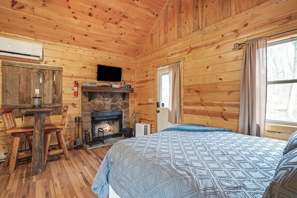 Ein gemütliches Blockhäuschen mit Kamin aus Stein, Holzwänden, einem Bett mit gemusterten Textilien, einem Fernseher an der Wand und einem Fenster mit Vorhängen.