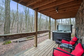 A covered outdoor area with a hot tub, wooden deck, and red cushioned chairs. - Hide-A-Way Tiny House | Hocking Hills, Hot Tub (Logan)