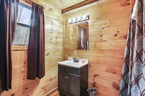 A wooden cabin bathroom with a sink, mirror, and window with curtains.