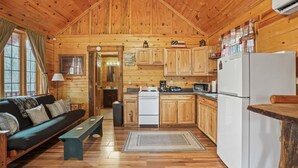 A cabin-style kitchen with wooden walls and floors, a stove, refrigerator, and a dining area with a bench. - Hide-A-Way Tiny House | Hocking Hills, Hot Tub (Logan)