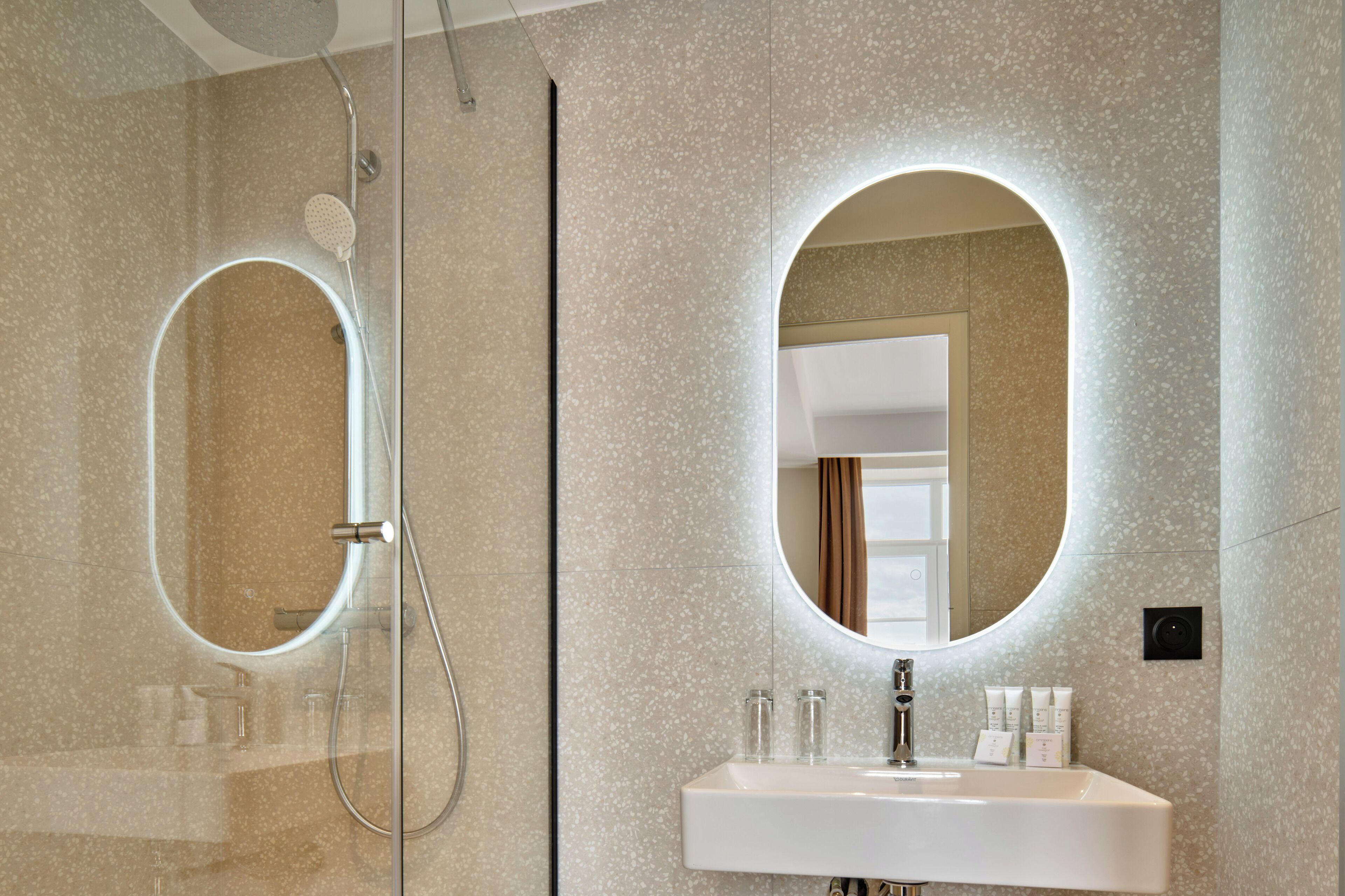 A modern bathroom with a glass-enclosed shower, a large oval mirror, and a white sink with toiletries.