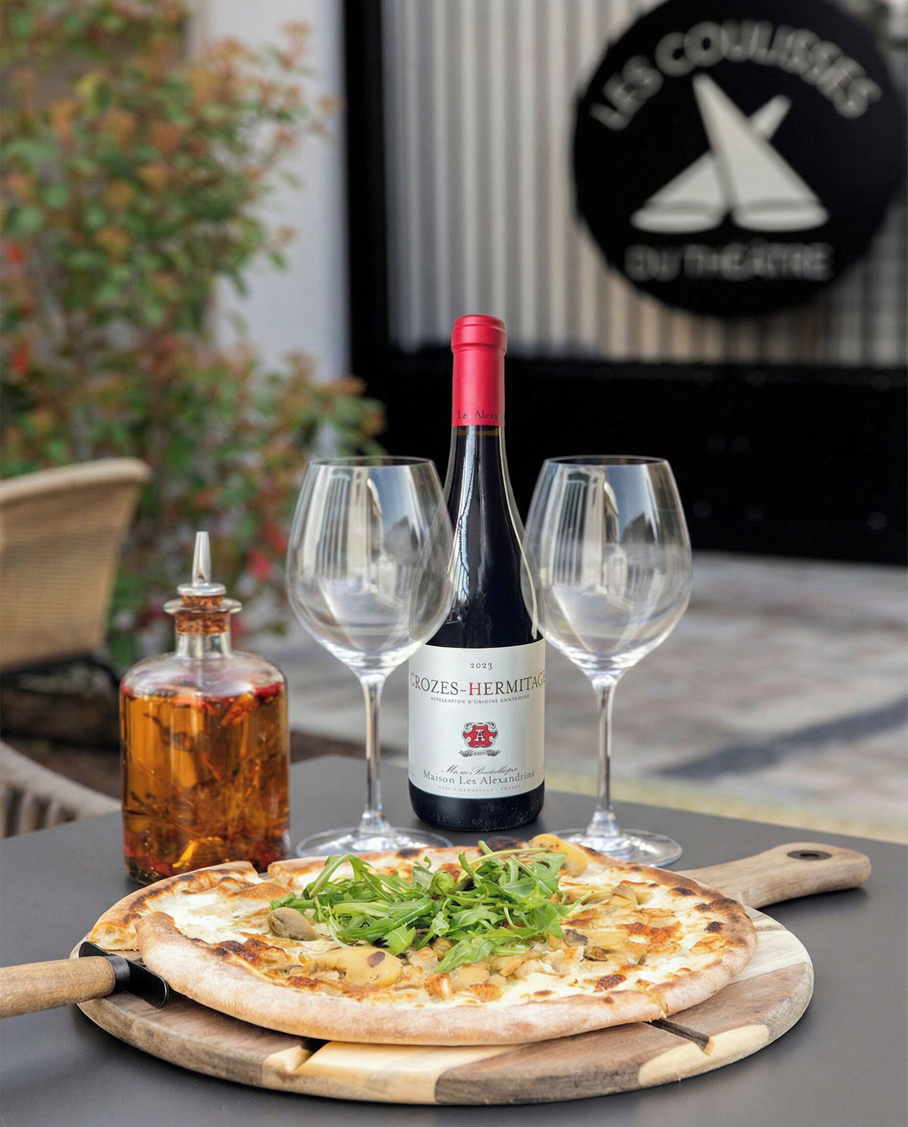 Een pizza op een houten plank, een fles Crozes-Hermitage wijn, en twee lege wijnglazen op een tafel.