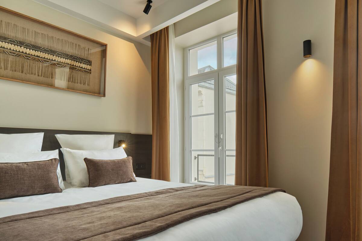 Une chambre d’hôtel avec un grand lit, une œuvre d’art encadrée au-dessus du lit, une fenêtre avec des rideaux et un luminaire fixé au mur.