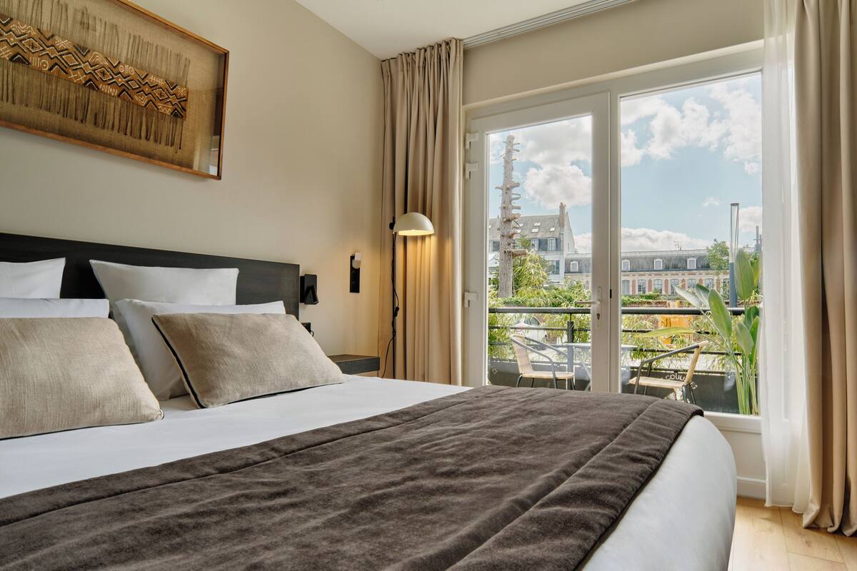 Une chambre d’hôtel avec un grand lit, une vue sur la ville depuis un balcon et une œuvre d’art encadrée au mur.