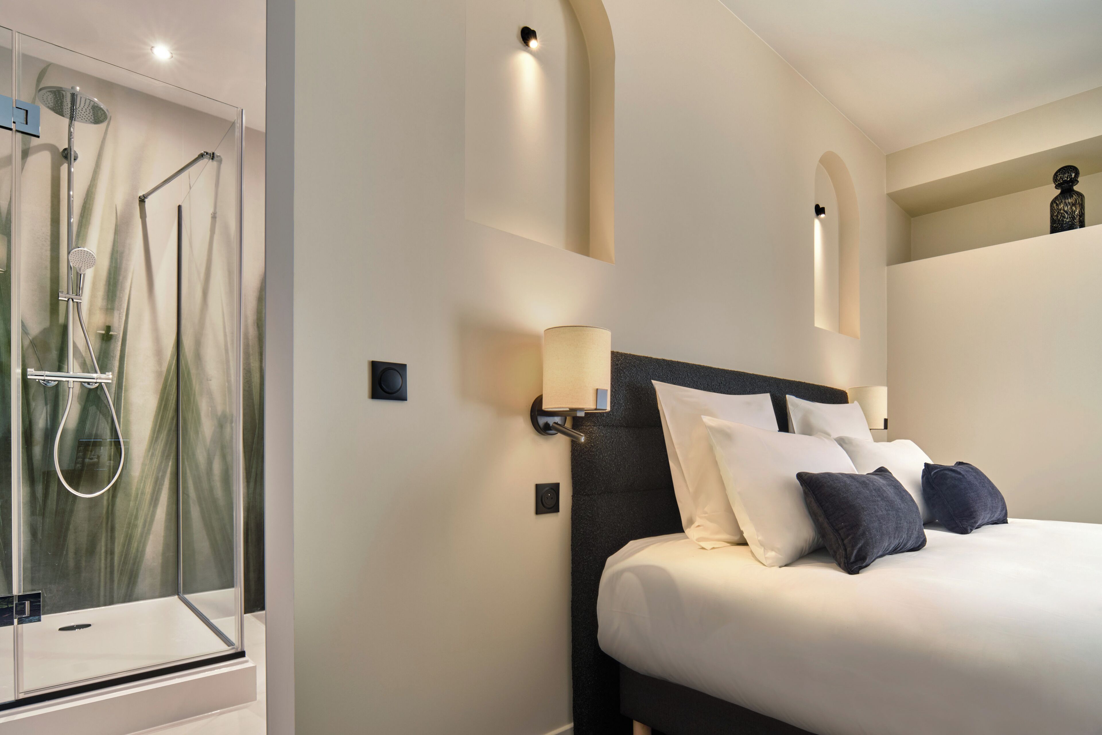 A hotel room with a glass-enclosed shower, a bed with white and dark pillows, and wall-mounted sconces.