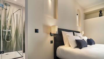A hotel room with a glass-enclosed shower, a bed with white and dark pillows, and wall-mounted sconces.