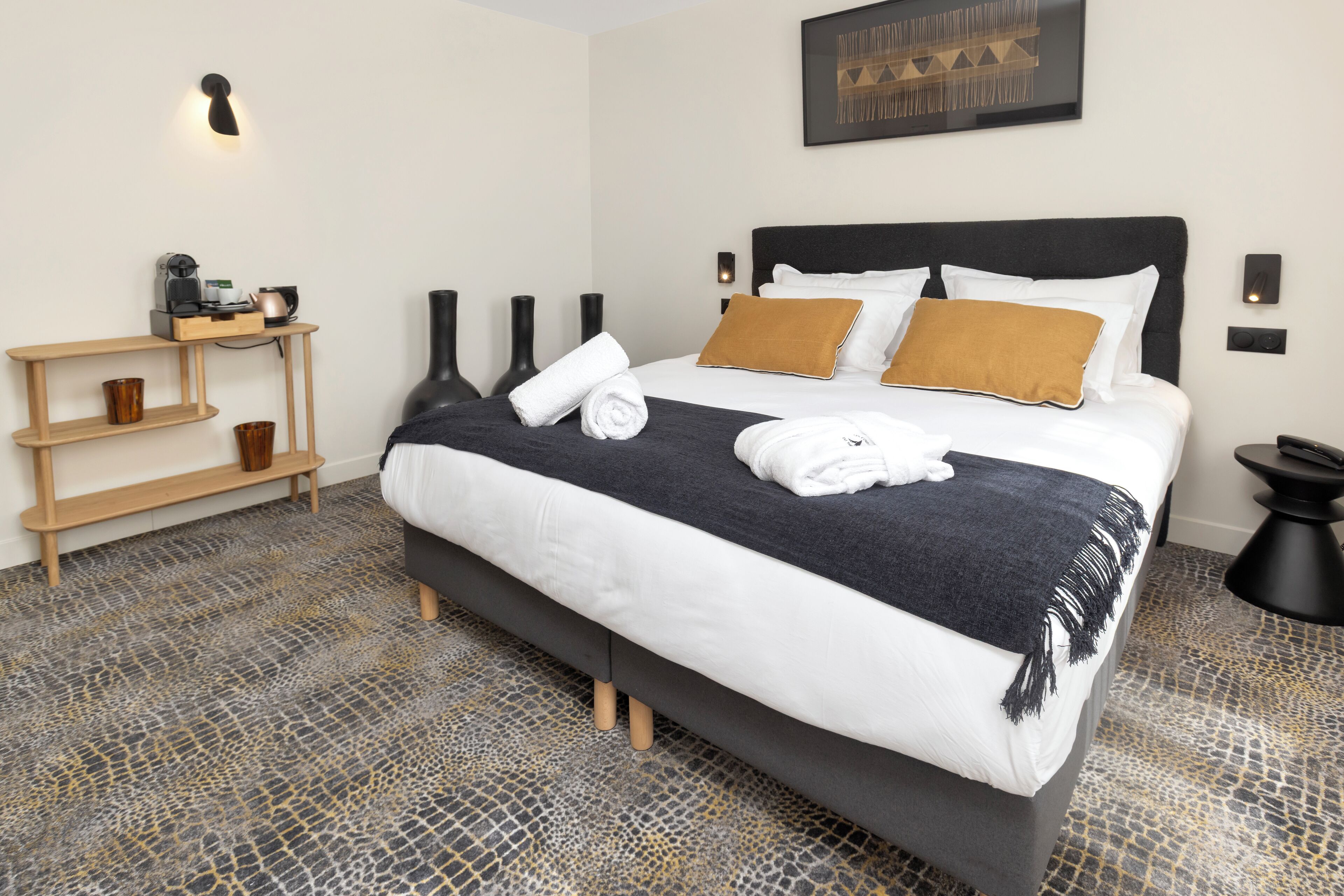 A modern hotel room with a large bed, a bedside table with a lamp, and a wall-mounted artwork.