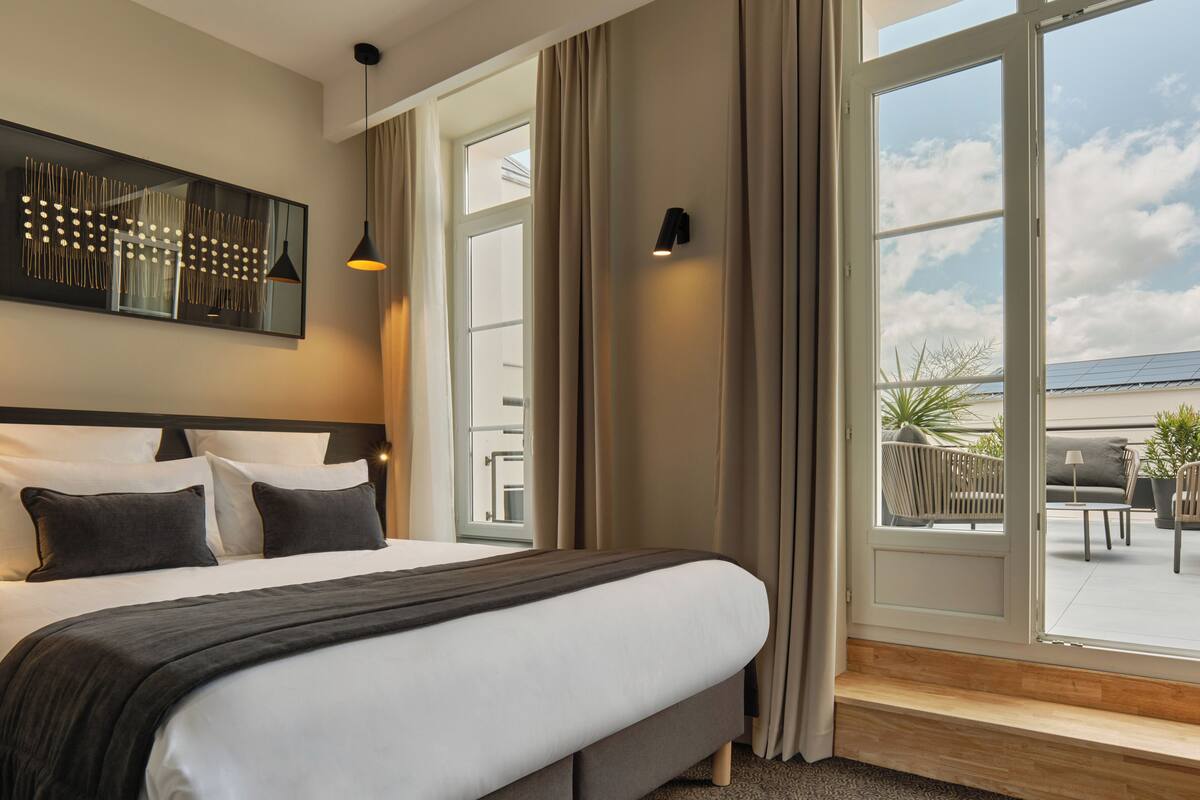 Une chambre d’hôtel moderne avec un grand lit, une vue sur l’espace extérieur aménagé en salon et une grande fenêtre avec des rideaux.