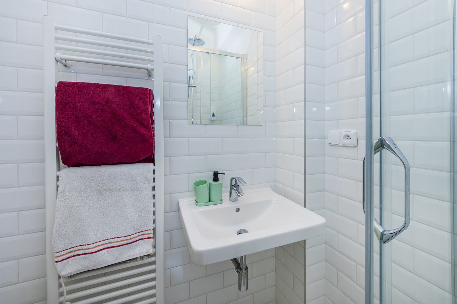 Une salle de bain avec des murs recouverts de carreaux blancs, un lavabo blanc avec un distributeur de savon vert, une cabine de douche en verre et deux serviettes sur un porte-serviettes blanc.