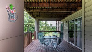 A covered patio with a metal table and chairs, a wooden deck, and a view of greenery.