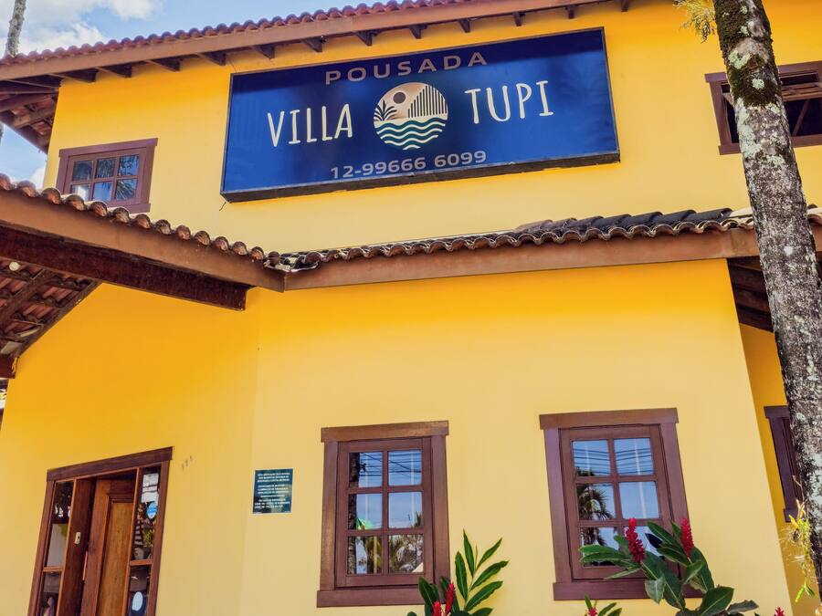A yellow building with a sign reading "Pousada Villa Tupi" and a phone number. There are windows and a door visible.