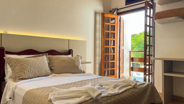 Un lit bien fait, avec une tête de lit, une porte en bois dotée de panneaux de verre et une vue sur la verdure à l’extérieur.