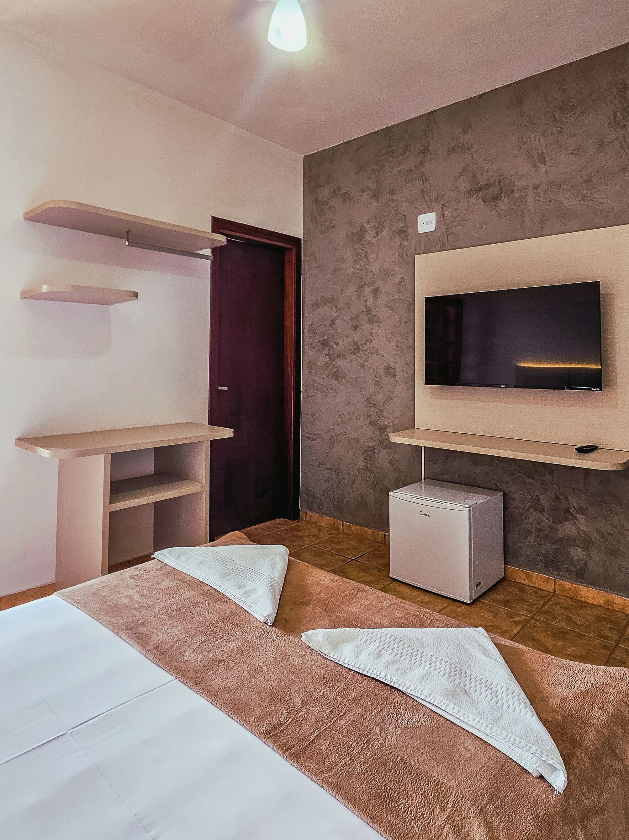 Quarto com uma cama, televisão fixada na parede, um pequeno armário e uma porta.