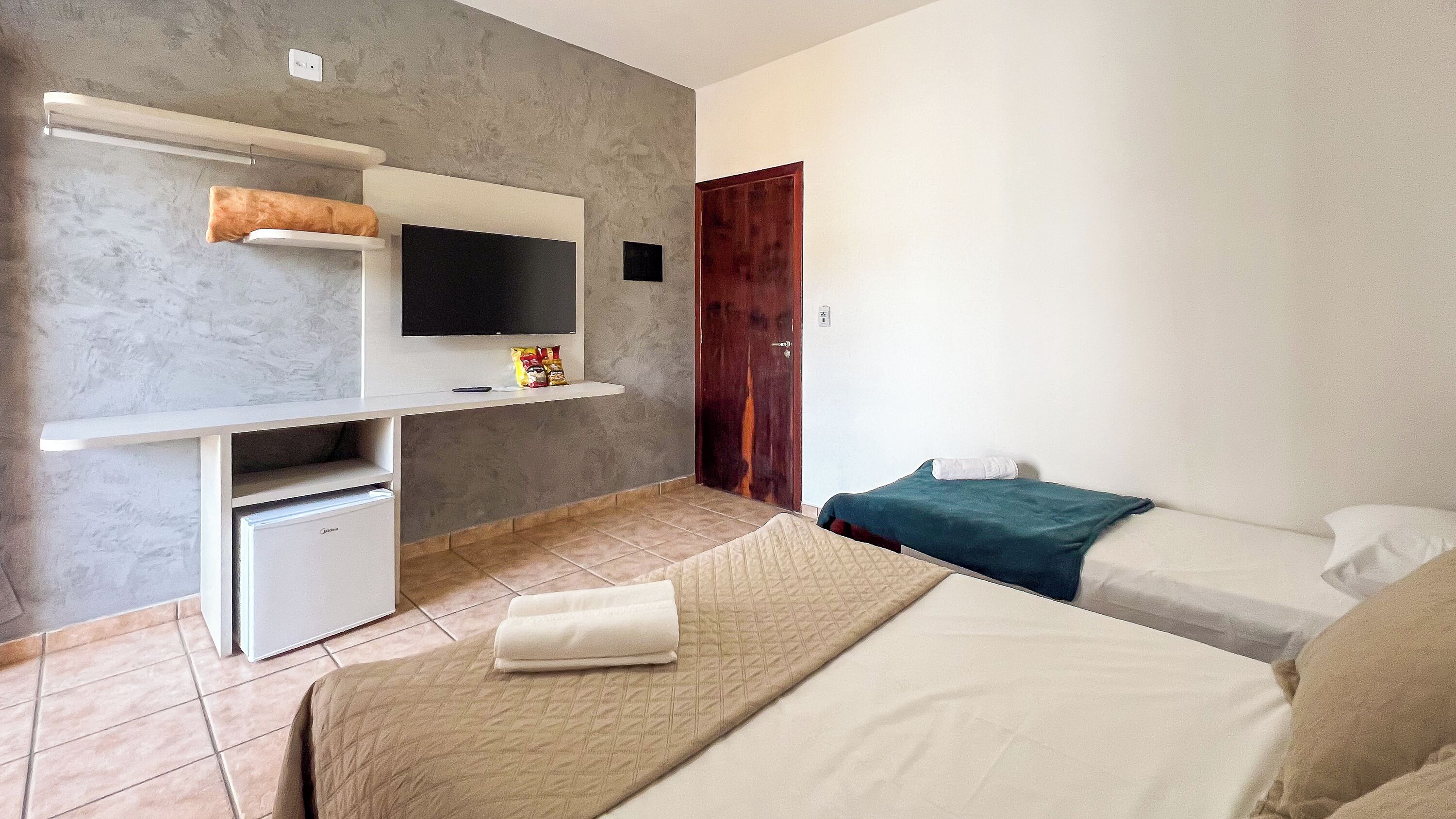 Een slaapkamer met een bed, een bureau met een televisie, een kleine koelkast en een houten deur.