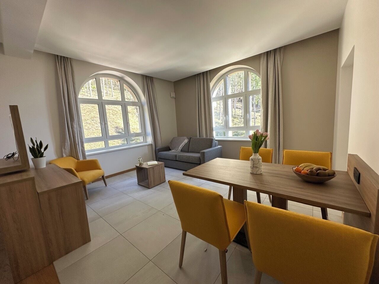 Un salon moderne avec un coin repas, comprenant un canapé, une table à manger avec des chaises jaunes et de grandes fenêtres avec des rideaux.