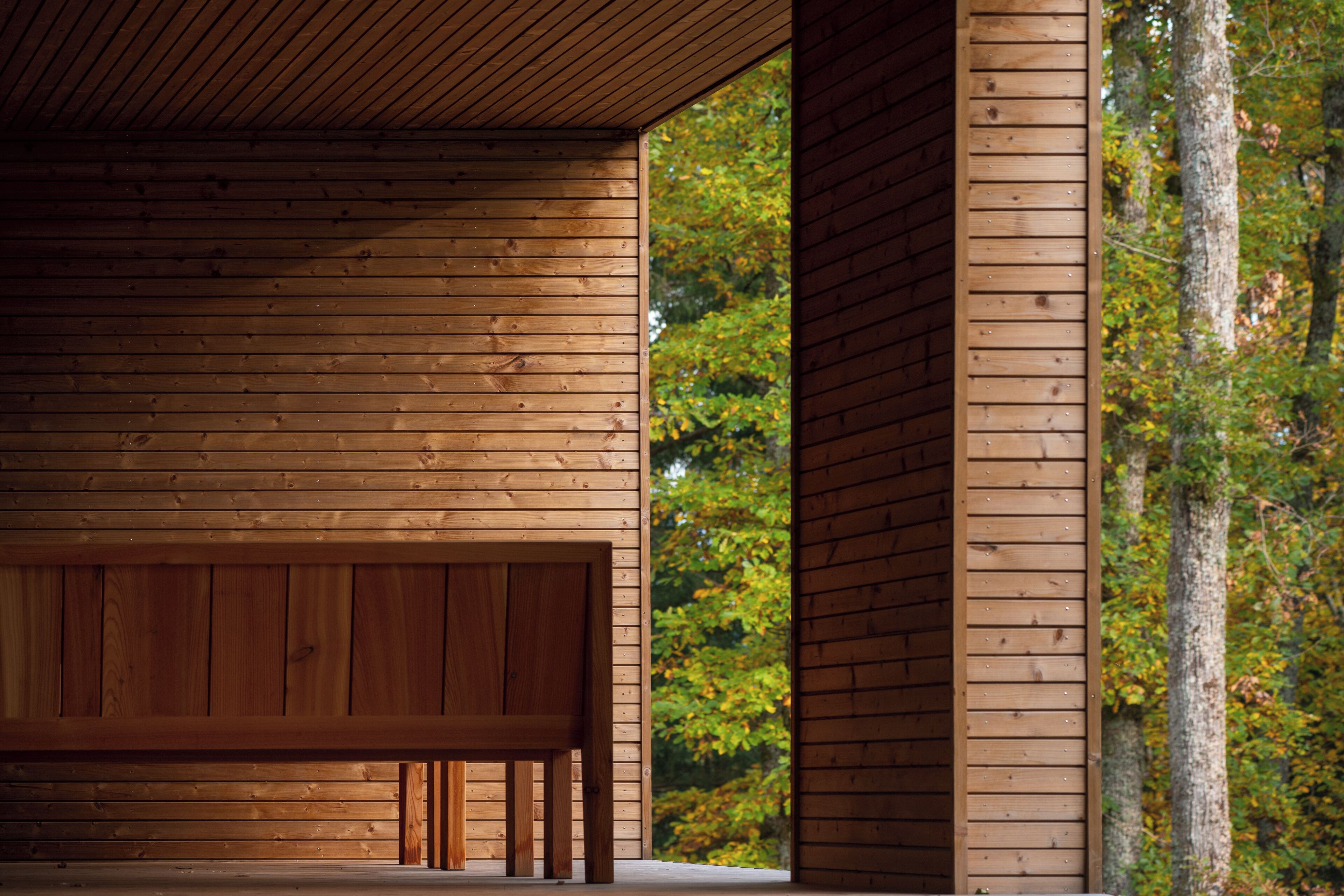 A wooden interior with a bench and a large window overlooking a forest.