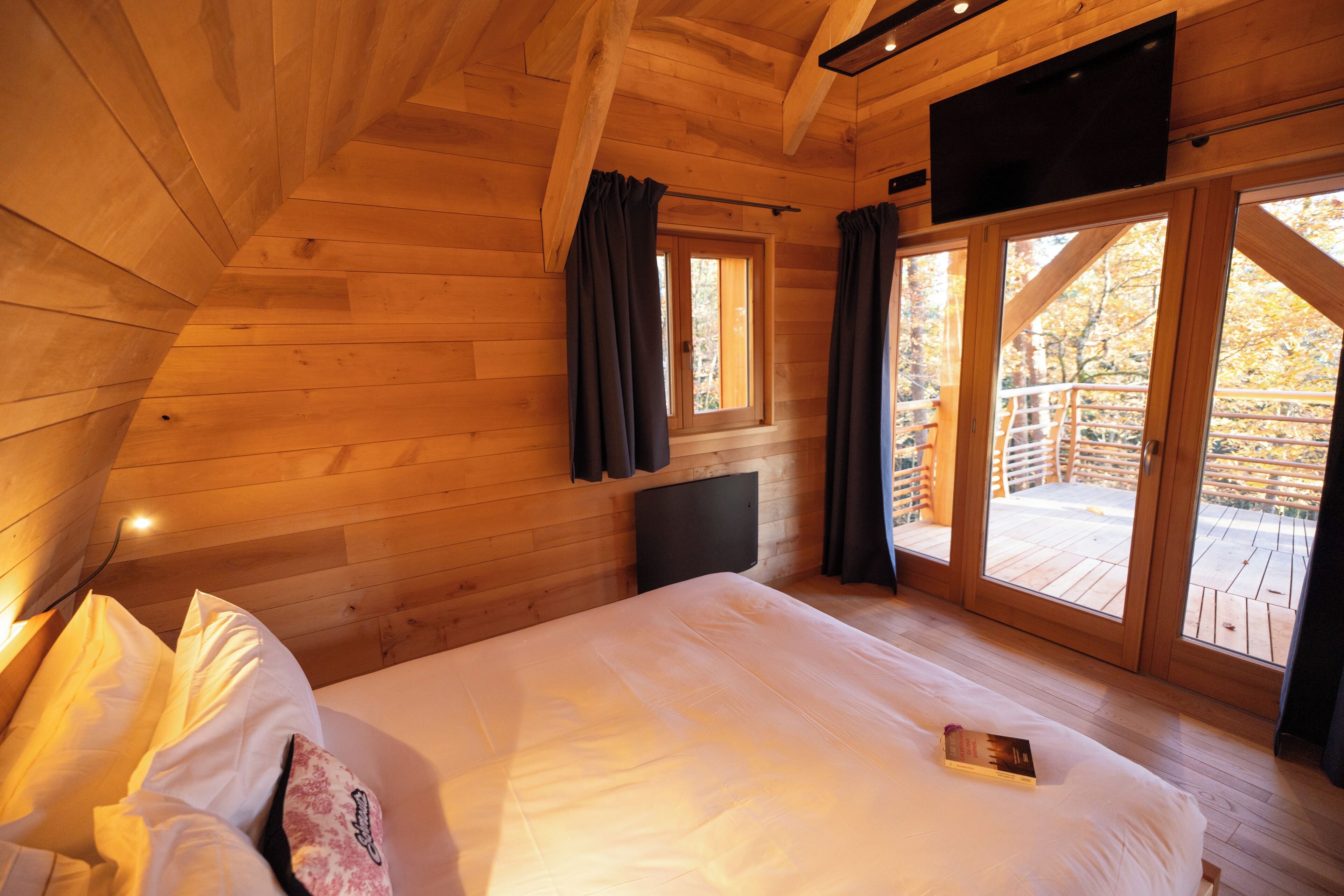 Een houten slaapkamer met een bed, een aan de muur bevestigde televisie en een raam met gordijnen.