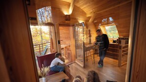 A cozy cabin interior with wooden walls, a dining area, and a large window offering a view of trees. - Domaine de Haslach, The Originals Collection, Résidence de Tourisme (Munster)