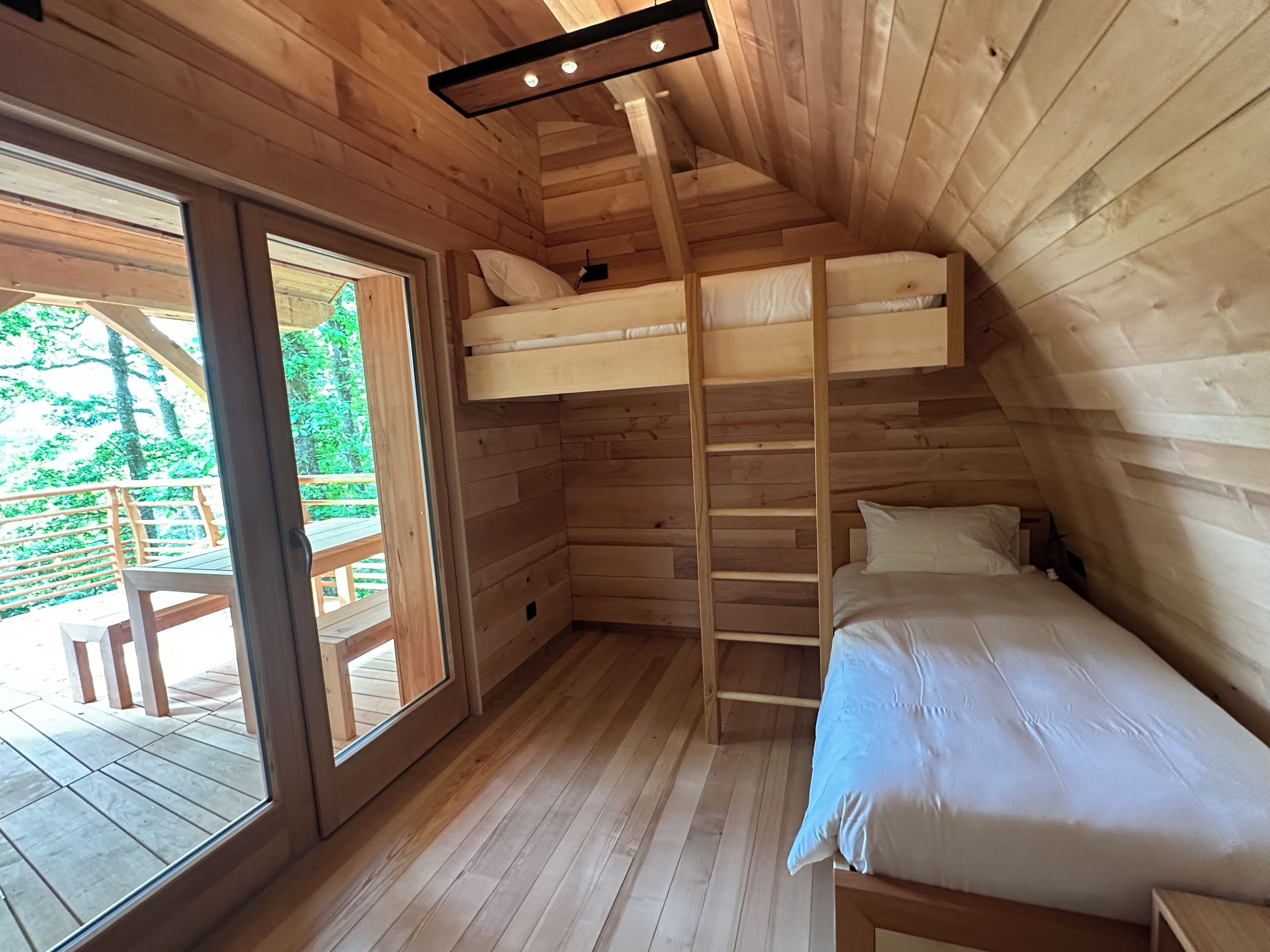 A wooden cabin room with a bunk bed, a ladder, and a large window.