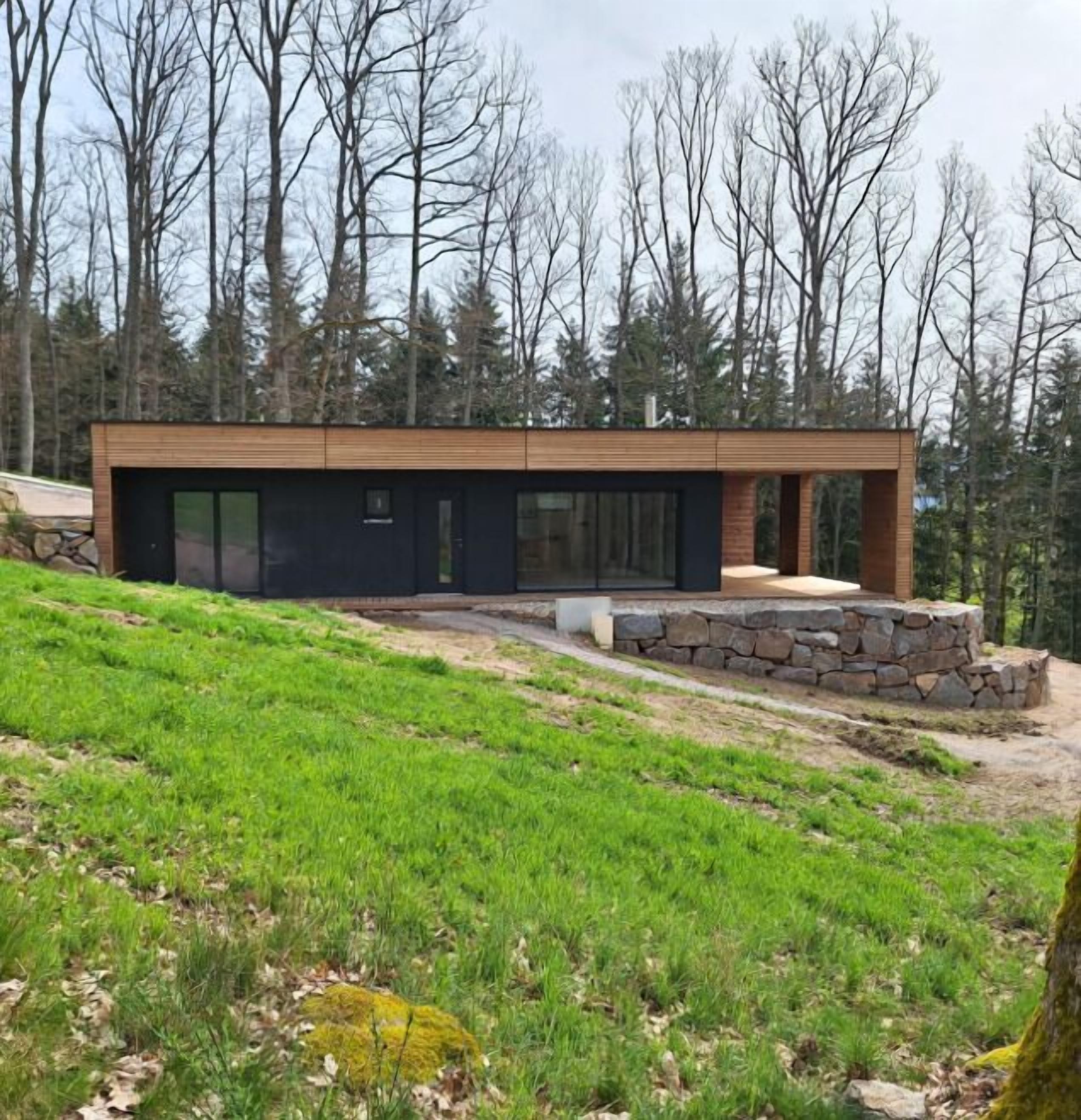 A modern two-story house with a stone foundation and wooden siding, surrounded by a grassy landscape and trees.