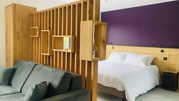 Una habitación de hotel moderna con un separador de madera, un sofá gris, una cama con ropa de cama blanca y una pared de acento morada.