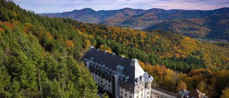 A large, historic building with a symmetrical facade, surrounded by dense forest and mountains in the background.