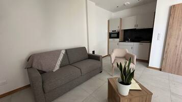Una sala de estar moderna con un sofá gris, una mesa de centro de madera con una planta, y una cocina al fondo.