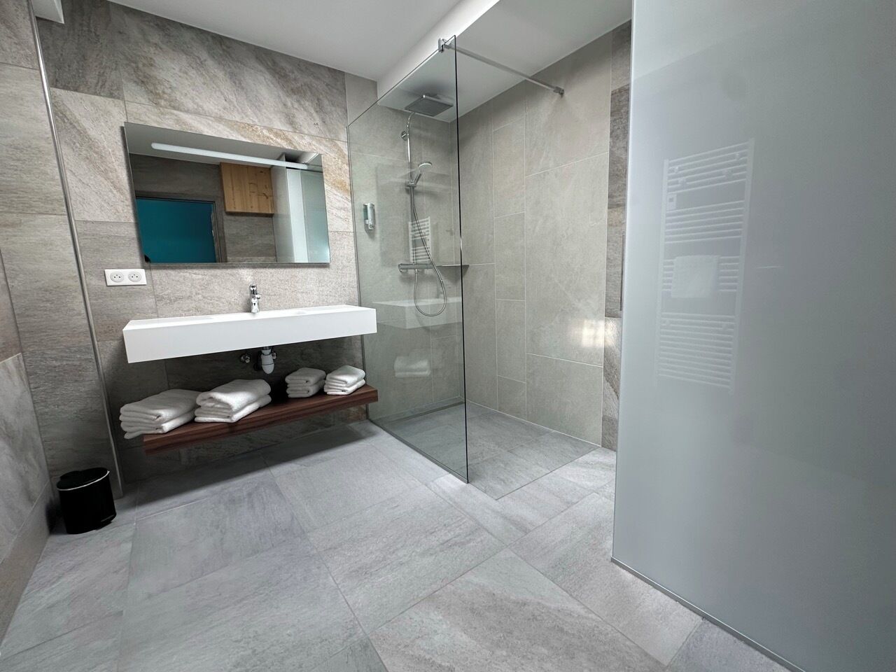 Une salle de bain moderne avec une douche à l’italienne en verre, un lavabo blanc doté d’une étagère en bois et un miroir au-dessus du lavabo.