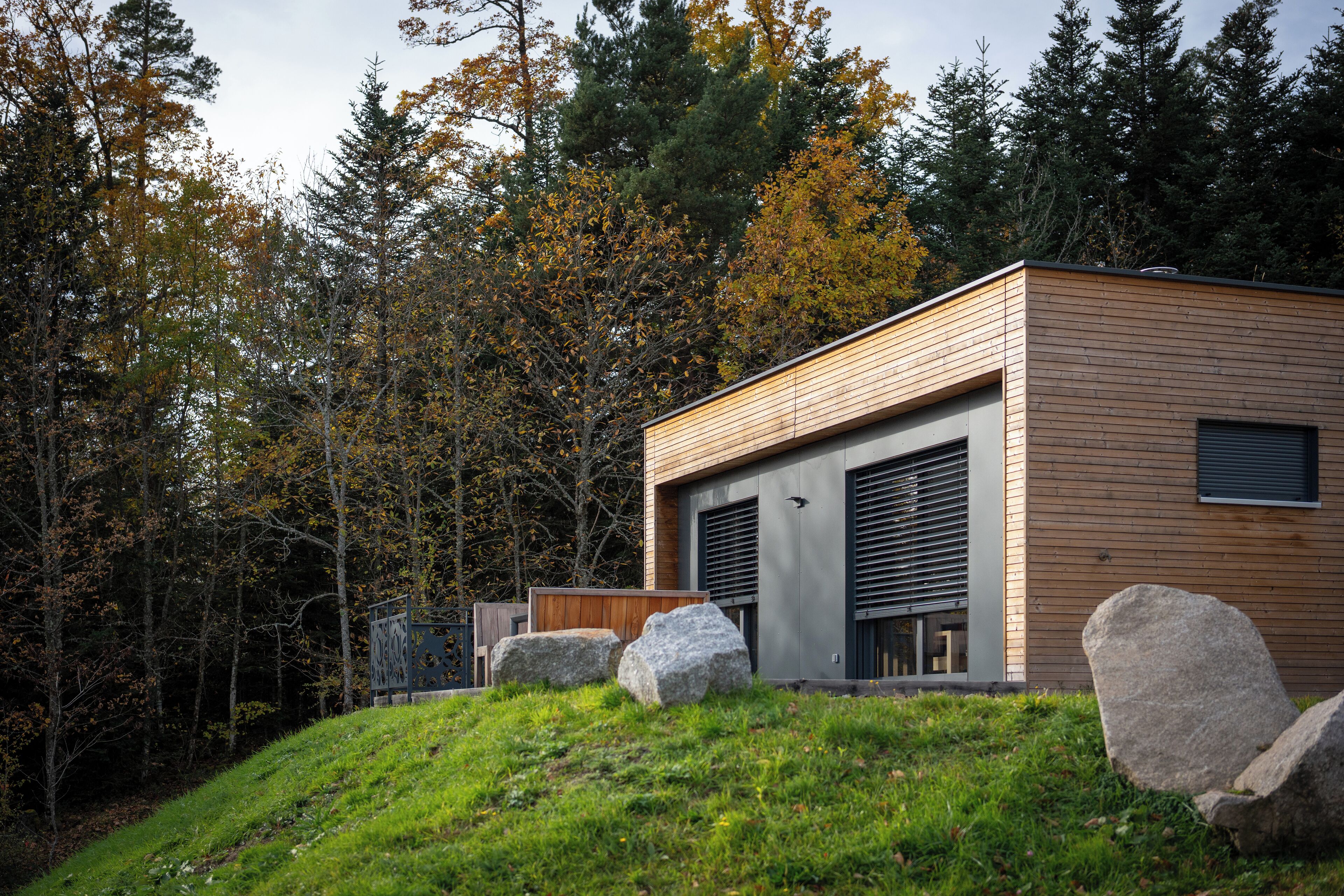 Un bâtiment moderne avec une façade en bois, de grandes portes de garage et une colline verdoyante parsemée de rochers.