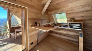 A wooden cabin with a kitchen area featuring a large window, a built-in oven, and a dining table. - Domaine de Haslach, The Originals Collection, Résidence de Tourisme (Munster)