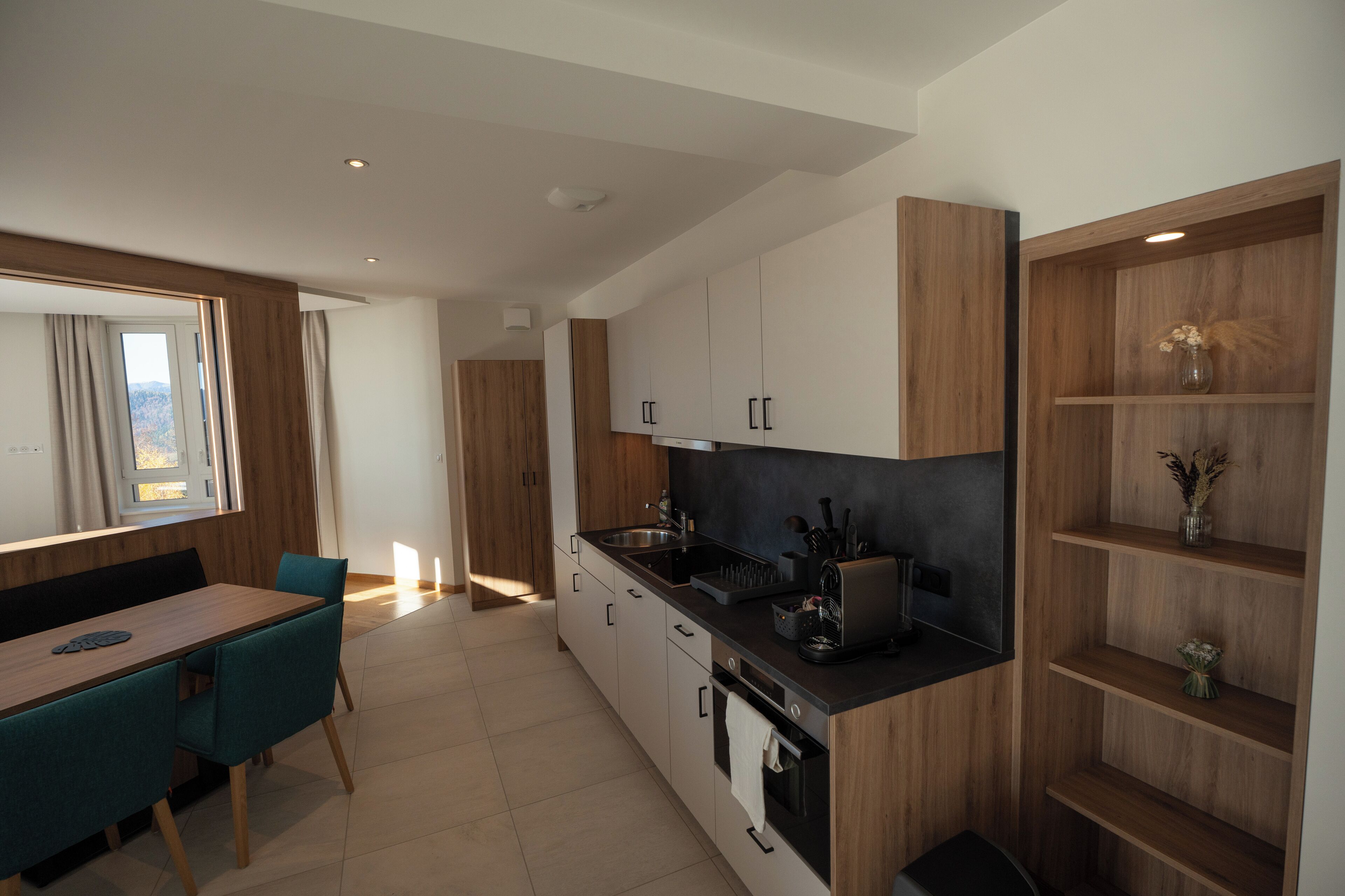 A modern kitchen with a dining area, wooden cabinets, and a built-in shelf with decorative items.