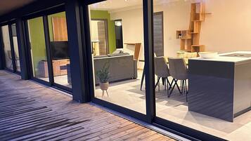 A modern interior with a wooden deck, large sliding glass doors, a kitchen area with a central island, and a dining space with chairs and a table.